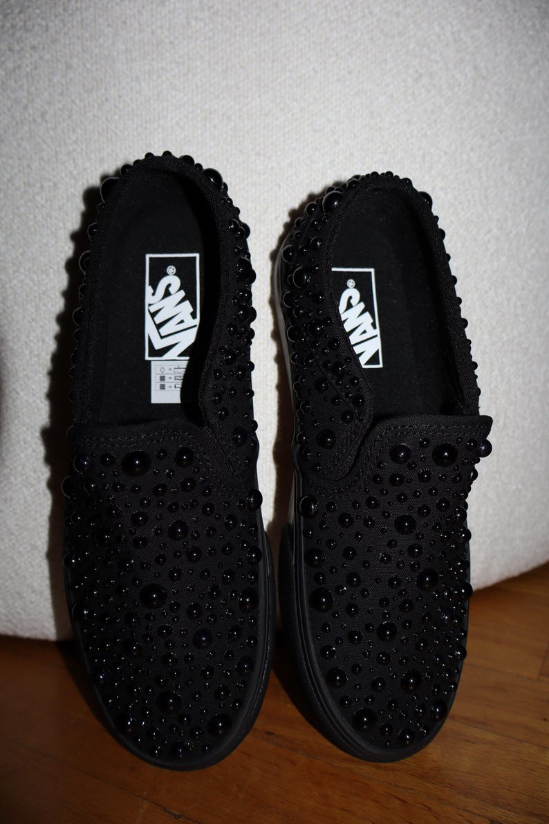 Puede incluir: Par de zapatillas negras sin cordones con un dise&ntilde;o tachonado. Los zapatos presentan el logotipo de Vans en blanco en el interior. Las zapatillas se exhiben sobre una superficie de madera.