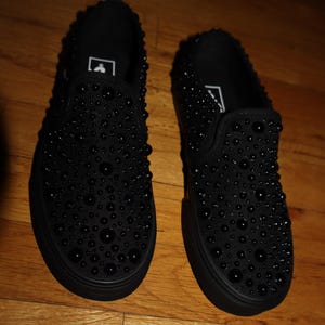 Puede incluir: Un par de zapatillas negras sin cordones adornadas con numerosos adornos negros y redondeados. Los zapatos tienen una suela negra s&oacute;lida y un dise&ntilde;o simple y minimalista. Las zapatillas se exhiben sobre una superficie de madera.
