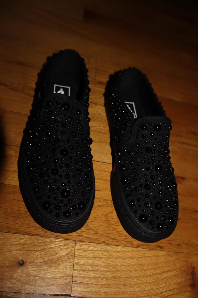 Puede incluir: Un par de zapatillas negras sin cordones adornadas con numerosos adornos negros y redondeados. Los zapatos tienen una suela negra s&oacute;lida y un dise&ntilde;o simple y minimalista. Las zapatillas se exhiben sobre una superficie de madera.