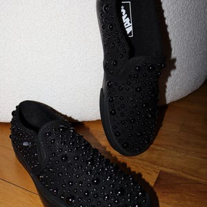 Puede incluir: Zapatillas negras sin cordones cubiertas de peque&ntilde;os adornos redondos negros. Los zapatos tienen una suela negra y el logotipo de Vans es visible en el interior. Los zapatos se exhiben sobre un suelo de madera.