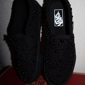 Puede incluir: Par de zapatillas negras sin cordones cubiertas de peque&ntilde;os adornos redondos y oscuros. Los zapatos tienen suela negra y el logo de Vans es visible en la plantilla. Los zapatos se exhiben encima de una caja de zapatos roja y marr&oacute;n.