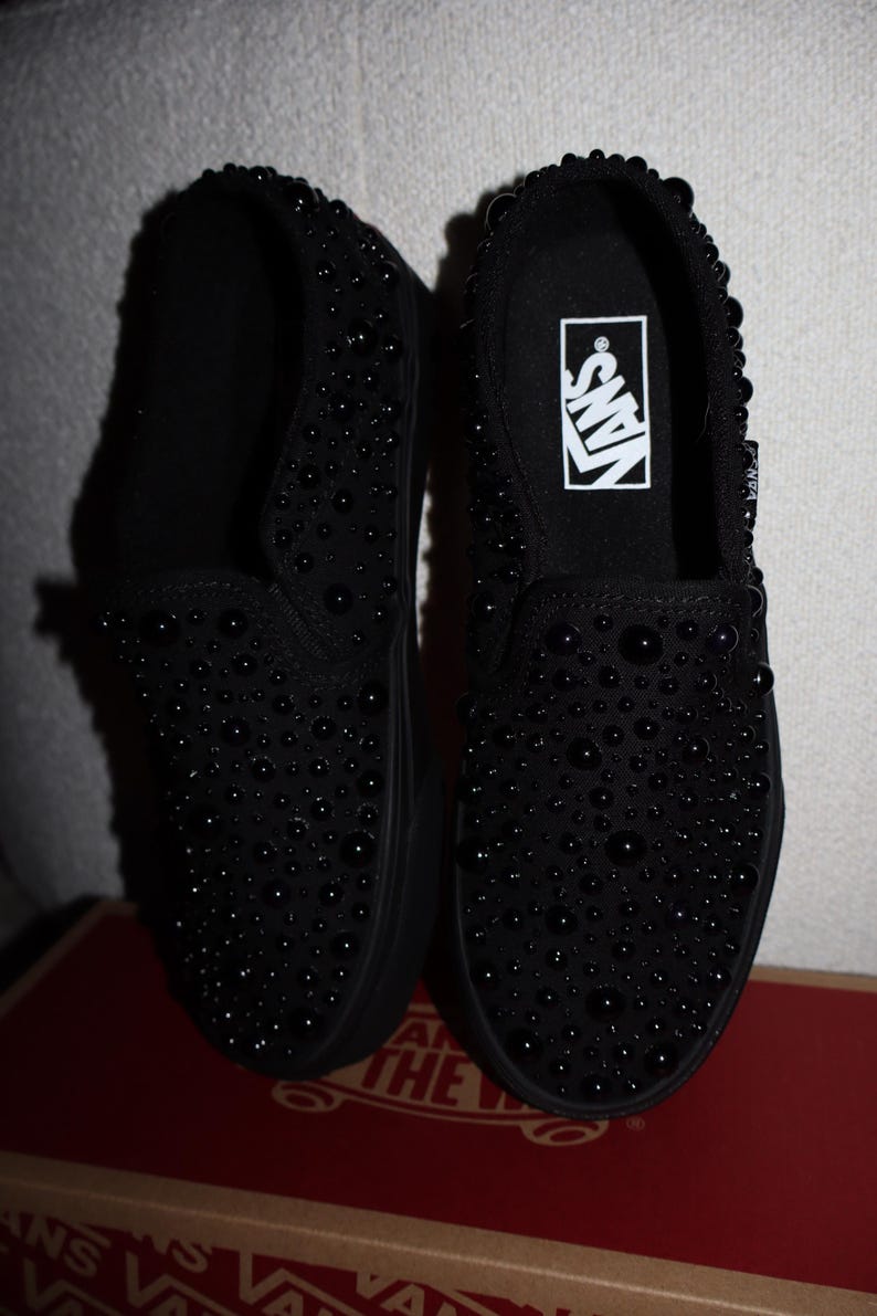 Puede incluir: Par de zapatillas negras sin cordones cubiertas de peque&ntilde;os adornos redondos y oscuros. Los zapatos tienen suela negra y el logo de Vans es visible en la plantilla. Los zapatos se exhiben encima de una caja de zapatos roja y marr&oacute;n.