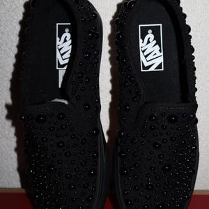 Puede incluir: Un par de zapatillas negras sin cordones cubiertas de peque&ntilde;os adornos negros redondos. Las zapatillas tienen una suela negra y el logotipo de Vans en blanco en la plantilla. Los zapatos se muestran en una caja roja y blanca.