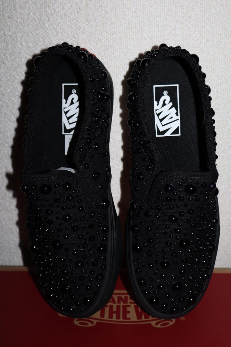 Puede incluir: Un par de zapatillas negras sin cordones cubiertas de peque&ntilde;os adornos negros redondos. Las zapatillas tienen una suela negra y el logotipo de Vans en blanco en la plantilla. Los zapatos se muestran en una caja roja y blanca.