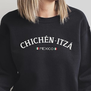 May include: A black long-sleeve crewneck sweatshirt featuring the white text CHICHÉN-ITZÁ in an arched font. Below the text is the word MEXICO in a smaller font, flanked by two small horizontal icons of the Mexican flag in green, white, and red.