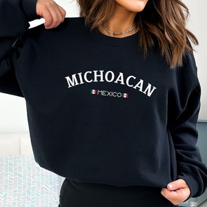 May include: A black long-sleeve crewneck sweatshirt featuring the white text MICHOACAN in an arched font. Below the text is the word MEXICO in smaller white letters, flanked by two small horizontal icons of the Mexican flag in green, white, and red.