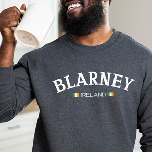 May include: Dark gray sweatshirt with the word "BLARNEY" in white arched text, with "IRELAND" below and the Irish flag. A white mug is held up in the air. The sweatshirt is a crew neck style.