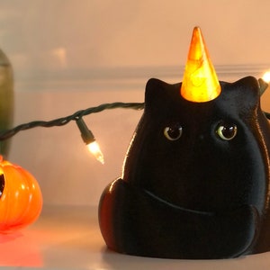 May include: A black cat figurine with a yellow cone hat, sitting on a white surface with a string of lights. The cat has large, round eyes and a small, pointed nose.