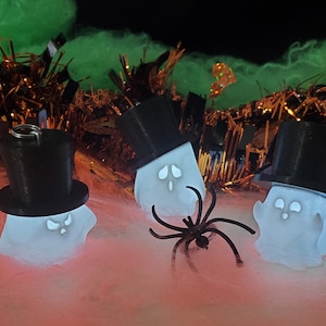 May include: Three white ghost ornaments with black top hats. The ghosts are glowing and have a spooky expression. The ghosts are sitting on a pink and white fluffy surface with a black spider in the foreground.