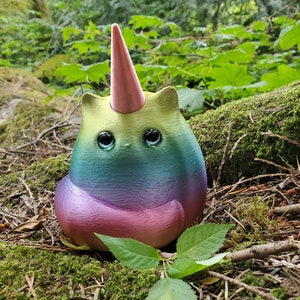 May include: A rainbow-colored cat figurine with a pink unicorn horn, sitting on a bed of moss and pine needles.