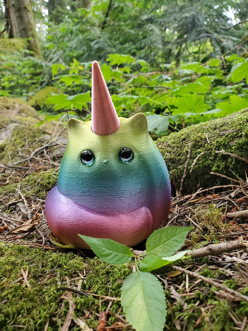 May include: A rainbow-colored cat figurine with a pink unicorn horn, sitting on a bed of moss and pine needles.