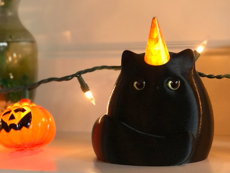 May include: A black cat figurine with a yellow cone hat, sitting on a white surface with a string of lights. The cat has large, round eyes and a small, pointed nose.