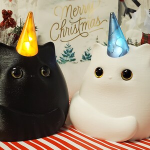May include: Two ceramic cat figurines with unicorn horns, one black and one white, wearing festive hats. The black cat has a yellow hat with a light inside, and the white cat has a blue hat with a swirling pattern.
