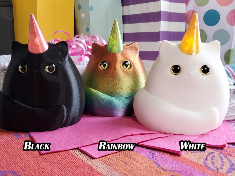 May include: Three ceramic cat figurines with unicorn horns and party hats. The cats are black, rainbow, and white.