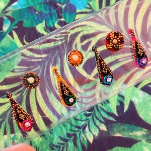 May include: A collection of decorative nail art stickers in various shapes and colors. The stickers include teardrop, round, and oval designs with gold, orange, pink, and blue accents. The stickers are displayed on a clear plastic sheet against a tropical leaf background.