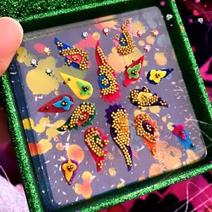 May include: A decorative box with a green glitter frame, holding colorful, abstract, beaded embellishments. The embellishments are in various shapes and colors, including red, yellow, blue, and purple, with gold bead accents. The background has a watercolor effect.