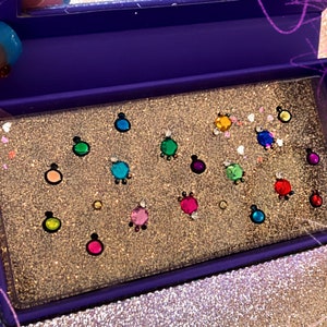 May include: A purple glittery makeup palette with a mirror. The palette contains a variety of colorful, jewel-like embellishments in shades of pink, green, blue, and yellow. The background has a glittery texture.