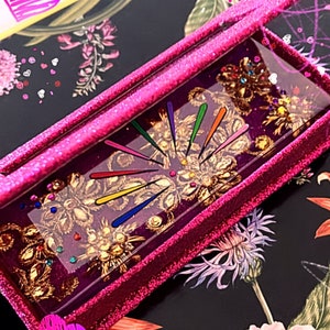 May include: A rectangular, glittery pink tray with a clear resin insert. The insert features gold floral embellishments, colorful lines radiating from a central point, and small, colorful gems. The tray sits on a floral patterned surface.