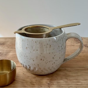 Könnte beinhalten: Eine gesprenkelte, weiße Keramiktasse mit einem goldenen Tee-Sieb. Die Tasse hat einen großen Henkel und eine strukturierte Oberfläche. Links von der Tasse befindet sich eine kleine goldene Schale. Ideal für Tee oder Kaffee.
