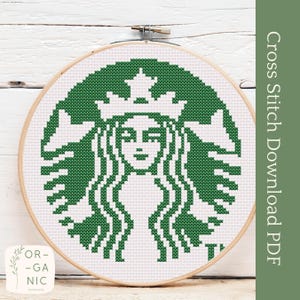 May include: Green and white cross stitch pattern of the Starbucks logo, featuring a mermaid with a crown and a star on her head. The pattern is displayed in a wooden embroidery hoop.