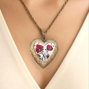 May include: Heart-shaped locket necklace with a floral design. The pendant features a white background with red flowers and green leaves. The locket is framed with a gold-colored filigree design and hangs from a delicate chain.
