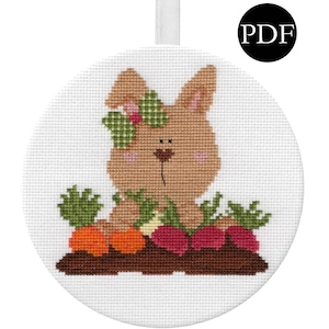 May include: A brown bunny with a green flower on its head sits in a patch of carrots and beets. The bunny is surrounded by green leaves. The image is a cross stitch pattern.