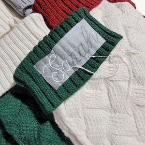 Könnte beinhalten: Ein grüner Strickstrumpf-Umschlag mit einem halbtransparenten Aufnäher, auf dem der Name Sarah in weißer Schreibschrift gestickt ist. Eine silberne Nähnadel mit einem weißen Faden liegt quer über dem gestickten Namen auf dem Stoff.