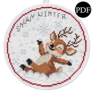 May include: A round, white cross-stitch ornament with a red border. The design features a smiling reindeer in brown, beige, and pink, with the words "Snowy Winter" stitched above. The reindeer is lying in the snow.