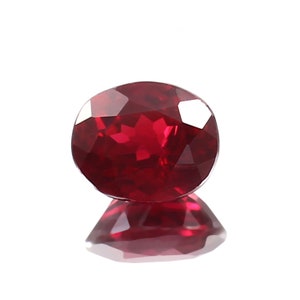 Puede incluir: Una piedra preciosa ovalada de color rojo oscuro con un corte facetado. La piedra preciosa refleja la luz y tiene un acabado brillante.