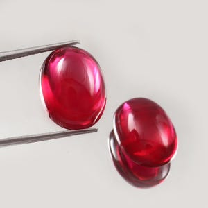Könnte beinhalten: Zwei ovale, polierte, tiefrote Edelsteine. Einer wird von einer Pinzette gehalten, der andere liegt auf einer reflektierenden Oberfläche. Die Edelsteine haben ein glänzendes, durchscheinendes Aussehen und zeigen Lichtbrechung.