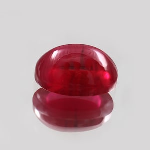 Pode incluir: Uma gema de rubi com corte cabochão, em forma oval. A pedra vermelha escura polida apresenta uma superfície refletora. A gema é apresentada sobre um fundo neutro, realçando sua cor e forma.