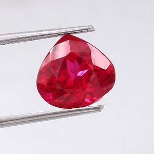 May include: A vibrant red pear-shaped gemstone with a faceted cut. The gemstone is held by a pair of tweezers.