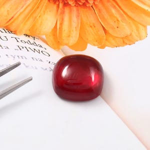 Op de afbeelding: Een gepolijste, vierkant geslepen, dieprode edelsteen. De edelsteen is glad en reflecterend, met afgeronde randen. De achtergrond omvat een bloem met oranje bloemblaadjes en een zilveren pincet.