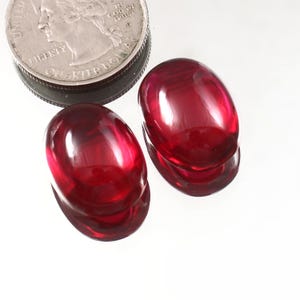 May include: Two oval-shaped, deep red gemstones with a high-gloss finish. A U.S. quarter coin is next to the gemstones for size comparison. The gemstones are on a white surface.