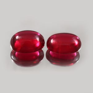 Peut inclure: Deux pierres précieuses ovales polies, d'un rouge profond. Les gemmes sont très réfléchissantes, avec une surface brillante. Les pierres sont présentées sur une surface gris clair, avec leurs reflets visibles en dessous.