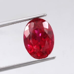 Könnte beinhalten: Ein leuchtend roter ovaler Edelstein, der von einer Pinzette gehalten wird. Der Edelstein ist facettiert und hochglanzpoliert.