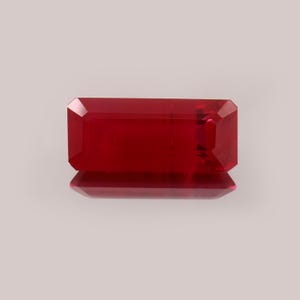 May include: A rectangular, faceted, deep red gemstone. The gem is highly polished, reflecting light and creating a brilliant appearance. The shape is an emerald cut, with a flat top and angled facets. The gem is set against a neutral background.