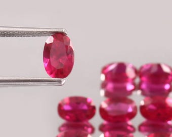 以下が含まれることがあります： ピンセットで保持された鮮やかな楕円形のルビー。深紅の色とカットされた表面が特徴です。他のルビーも複数あり、光を反射しています。宝石はニュートラルな背景に配置されています。