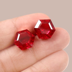Puede incluir: Dos piedras preciosas hexagonales rojas, probablemente rubíes, se muestran en una mano. Las piedras preciosas son facetadas y tienen un brillo brillante.