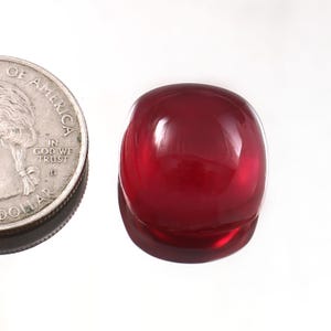 May include: A polished, deep red gemstone with a rounded, rectangular shape. The gem reflects light, showcasing its smooth surface. A US quarter coin is next to the gem for size comparison.