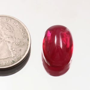Könnte beinhalten: Ein oval geschliffener, tiefroter Edelstein. Eine Münze dient als Größenvergleich. Die glänzende Oberfläche des Steins reflektiert das Licht und zeigt seine satte Farbe und Klarheit. Der Edelstein ist wahrscheinlich ein Rubin.