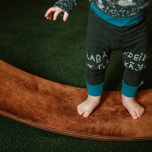 May include: A child stands on a wooden balance board. The child is wearing grey trousers with blue trim and white writing that says "LABA FI" on one leg and "KREIZ KAY" on the other leg.