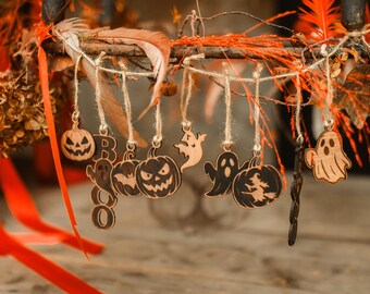 Puede incluir: Decoración de Halloween con adornos de madera con forma de calabazas, fantasmas y la palabra "BOO". Los adornos están suspendidos de una rama con hojas naranjas y marrones. El conjunto tiene un tema otoñal y espeluznante.