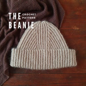 Könnte beinhalten: Eine beige Häkelmütze mit gerippter Struktur. Die Mütze liegt auf einer braunen Holzoberfläche. Der Text "THE CROCHET PATTERN BEANIE" ist über der Mütze sichtbar.