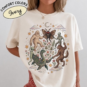 Può includere: Una t-shirt color avorio con una stampa grafica raffigurante un Bigfoot, una creatura simile a una falena con occhi rossi, uno scheletro, un alieno, un mostro marino e un lupo mannaro che si tengono per mano in cerchio tra stelle e fiori.