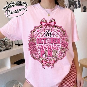 Op de afbeelding: Een lichtroze t-shirt met een grafisch ontwerp met de tekst In OCTOBER We Wear PINK. Het ontwerp bevat roze rozen, luipaardprint accenten, een lint voor borstkankerbewustzijn en roze-witte geruite strikken in een cirkelvormig kader.