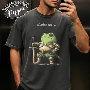 May include: A charcoal gray short-sleeve t-shirt featuring a graphic of a green frog dressed in golden armor and holding a sword. Above the frog, the text reads alpha male in a white typewriter-style font. The shirt has a crew neckline.