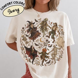 May include: An ivory t-shirt featuring a circular graphic of various Halloween figures including a witch, skeleton, werewolf, ghost, mummy, vampire, and pumpkin-headed character holding hands, surrounded by botanical elements and moths.