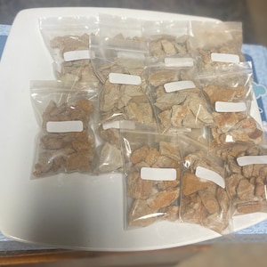 May include: Multiple small bags filled with light brown and tan rock-like pieces. The bags are arranged in rows on a white plate.