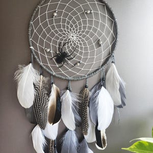 Könnte beinhalten: Ein grauer Traumfänger mit einem weißen Spinnennetz-Design und einer schwarzen Spinne in der Mitte. Der Traumfänger ist mit weißen, grauen und gemusterten Federn sowie kleinen Perlen verziert. Dieser dekorative Artikel ist perfekt für ein Schlaf- oder Wohnzimmer.